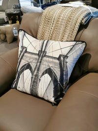 A PWFL1017 Bridge Pillow by Renwil, featuring a black and white bridge design, rests on a beige leather chair adorned with a knitted blanket, adding charm to the interior design crafted by our in-house artists.