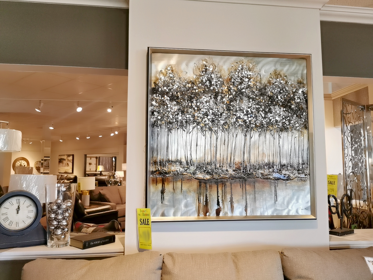 In the furniture store setting, the Metallic Forest Wall Art by Renwil, featuring an abstract painting of a tree line, hangs above a couch. The artwork showcases depth of color and is accentuated by eye-catching sale signs.