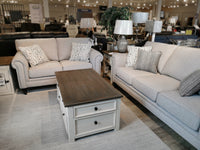 Showroom showcasing two beige Superstyle 9613 loveseats, a wooden coffee table, and lamps on side tables.