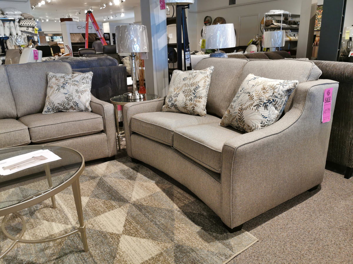 Explore our showroom showcasing the Superstyle 9670 Loveseat in gray upholstery, featuring medium-firm seating and adorned with floral patterned pillows. Complementing the stylish transitional design is a sleek glass coffee table along with floor lamps, crafting a distinctive living space.