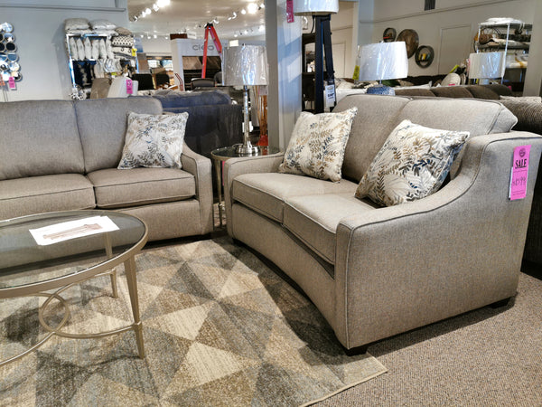 Explore a furniture showroom where a stylish transitional living space is highlighted by two Superstyle 9670 Loveseats, adorned with patterned pillows. Complementing the setup are a round glass coffee table and a geometric rug, all contributing to the elegant design.