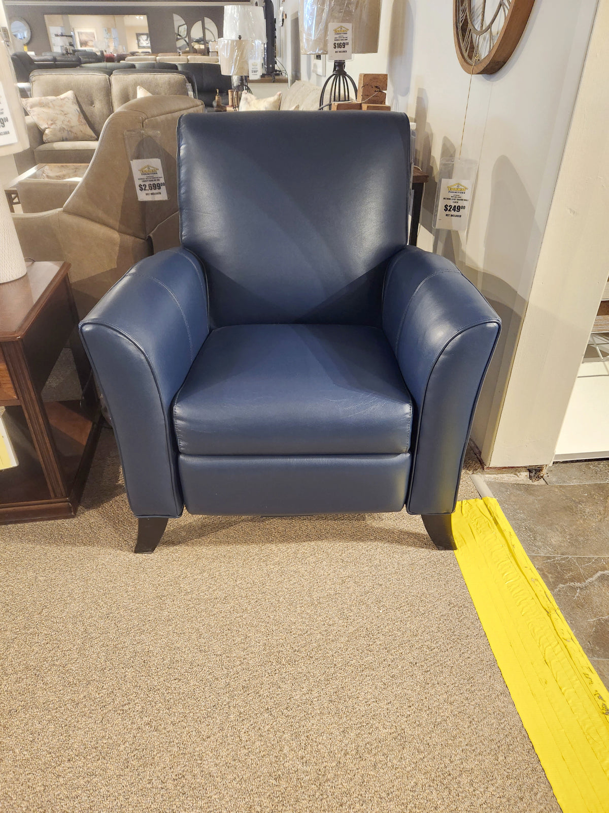 The L74 Push-Back Reclining Chair by Superstyle features a blue leather design with short wooden legs, displayed on a beige carpet in a furniture store. A price tag is visible on the side, highlighting its luxurious comfort.