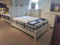 A bedroom showroom showcases the BB29552 Bonanza Bed by Vaughan Bassett, paired with a blue and white mattress, and accompanied by a nightstand and dresser made from hardwood solids, all arranged on a carpeted floor in transitional style.