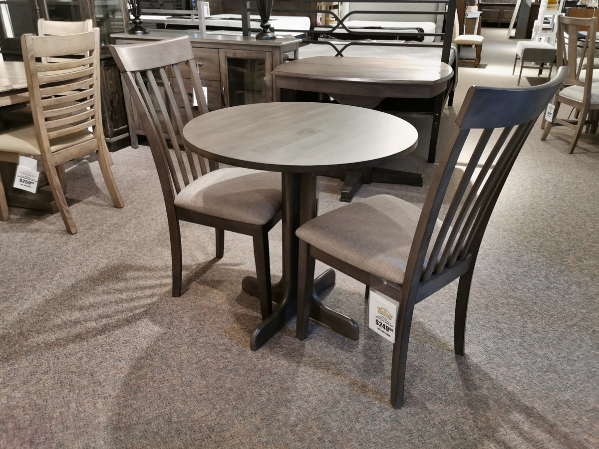 The DF30RD Delfini Dining Table Package by Winners Only is showcased in a furniture store showroom, featuring a small, round table crafted from solid hardwood with two matching chairs. It embodies transitional styling that seamlessly blends classic and modern aesthetics.