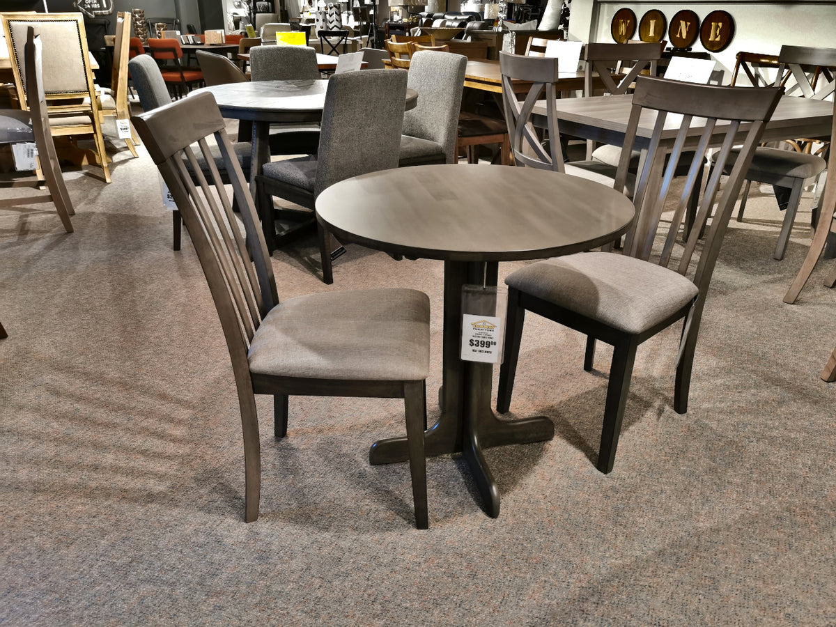 The DF30RD Delfini Dining Table Package by Winners Only features a small round table crafted from solid hardwood, paired with two wooden chairs upholstered in gray fabric, and is showcased in a furniture store to highlight its contemporary styling.