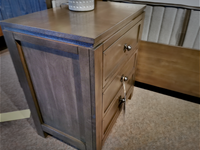 A stylish Lancaster Nightstand from Winners Only, made of wood with a grey/brown finish and part of the Lancaster bedroom collection, features three drawers and a ceramic pot on top. It is elegantly placed on a carpeted floor beside the bed.