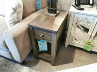 A contemporary side table made of birch wood, displaying a sale tag, is placed next to a couch and white cabinet. The Lancaster Lamp Table by Winners Only serves as a stand for both a lamp and phone.