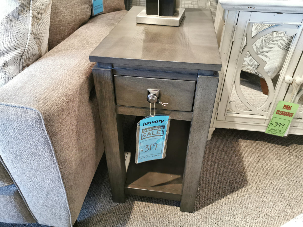 A Lancaster Lamp Table by Winners Only, featuring a drawer and tagged for sale, is positioned next to a couch in the furniture store's contemporary display, highlighting its elegant birch wood finish.