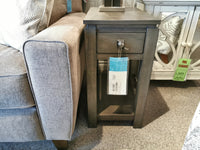 A wooden side table made from birch wood, featuring a drawer and shelf, perfectly complements the beige sofa. Nearby, the Winners Only Lancaster Lamp Table embodies contemporary design, while a white cabinet with a green tag adds a touch of contrast.