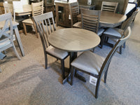 The store display features the DF30RD Delfini Dining Table Package by Winners Only, showcasing round wooden tables and chairs made from solid hardwood, all arranged on a carpeted floor in a transitional style.