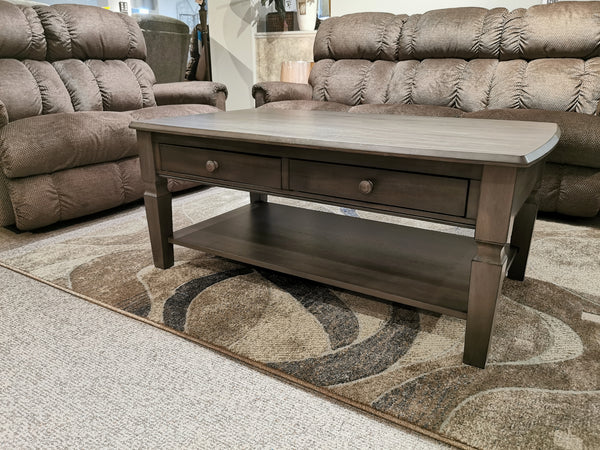 An Annapolis 48" Coffee Table by Winners Only, crafted with solid hardwood and featuring drawers, sits on a patterned rug, surrounded by brown upholstered sofas in the living room.