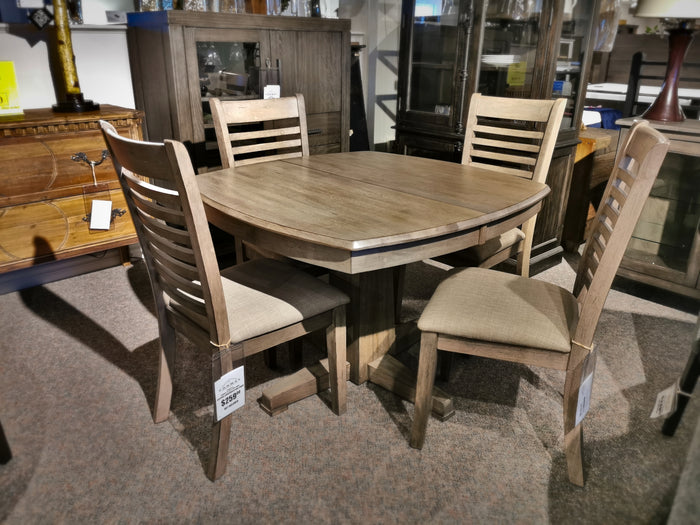 The Ventura Dining Package by Winners Only features a wooden dining set with a round table and four cushioned chairs, elegantly displayed in a furniture store. Crafted from solid hardwood, it boasts a chic grey washed finish that adds a touch of modern sophistication to any dining space.